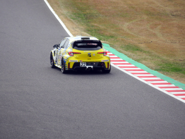 Ein gelber Sportwagen mit Text und Zahlen darauf, der eine Rennstrecke entlangfährt, die von Gras gesäumt ist.