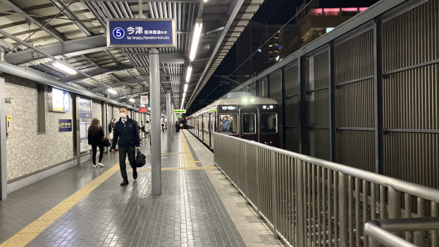 Ein Zug fährt auf ein Bahnsteiggelände zu, mit Menschen in der Nähe, einem Geländer auf der rechten Seite, Gebäuden im Hintergrund, Bahnsteigbeleuchtung und einer Anzeigetafel mit Text.