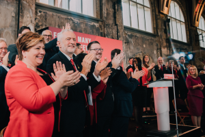 Eine Gruppe von Menschen, wahrscheinlich Liberale, klatscht feiernd vor einer Menge, mit einem Podium, Mikrofon und Texttafel rechts, Stühlen, einer Fahne, Wand, Fenstern und Lichtern im Hintergrund.