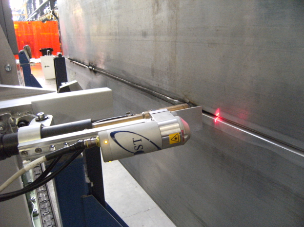 Hell leuchtendes Laserlicht, das aus einer Schneidemaschine in einer industriellen Fabrikumgebung austritt, mit einer Wand auf der rechten Seite und Hintergrundobjekten.