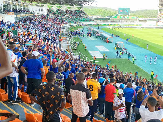 Eine große Menschenmenge versammelt sich vor einem eingezäunten Fußballfeld mit Hügeln und einem klaren Himmel und erwartet ein Spiel während der Afrikameisterschaft 2019.