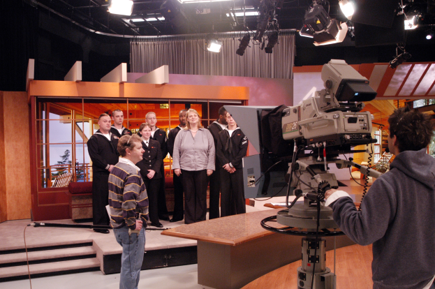 Gruppe von Menschen in einem Fernsehstudio mit einer Person, die eine Kamera hält, Lampen an der Decke sichtbar.