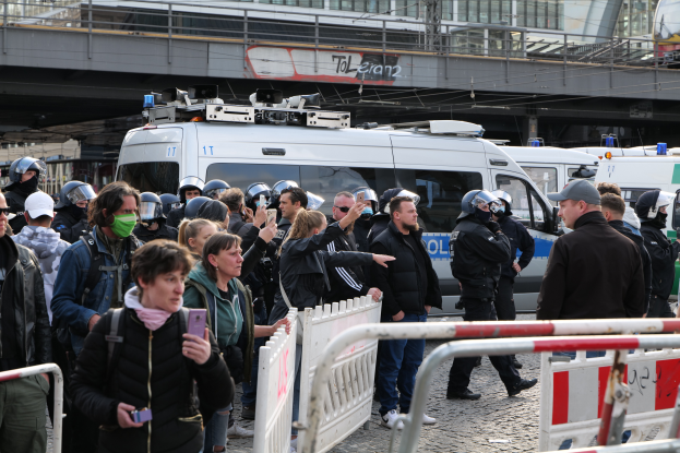 Eine Gruppe von Menschen steht vor einer Reihe von Polizeiwagen, einige tragen Helme und halten Telefone, mit Absperrungen im Vordergrund und einer Brücke mit Geländern und Gebäuden im Hintergrund in Berlin, Deutschland.