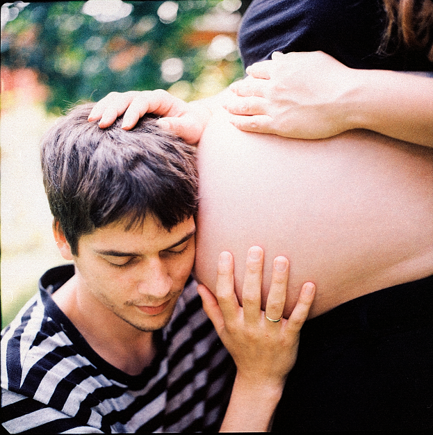 Ein Mann und eine Frau stehen nebeneinander, der Mann berürhrt zart den Bauch der Frau und schaut auf ihr ungeborenes Kind.