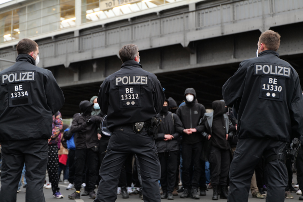 Polizeibeamte in schwarzer Uniform und Masken vor einer Menge stehend, mit einer Brücke und einem Gebäude im Hintergrund.