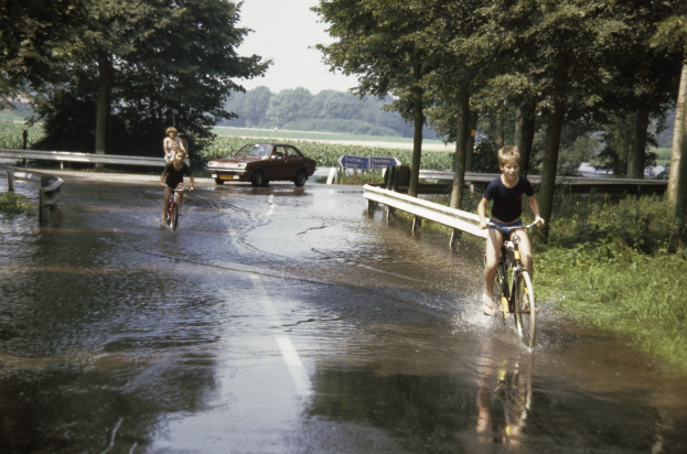 Eine Gruppe von Menschen, die auf Fahrrädern eine Überschwemmte Straße entlangfahren, mit einem danebenfahrenden Auto, gesäumt von Geländern und Vegetation unter einem klaren blauen Himmel.