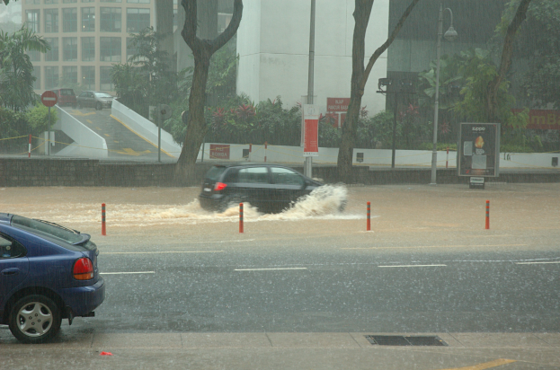 Ein Auto fährt durch eine überflutete Straße bei Regen, mit Wasser, das um das Auto spritzt, umgeben von Straßenschildern, Straßenschildern, Straßenlaternen, Bäumen, Pflanzen und Gebäuden mit Fenstern im Hintergrund.