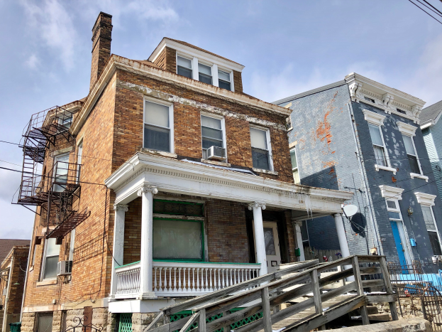 Eine Reihe heruntergekommener Häuser an einer Straßenecke, mit kaputten Fenstern, bröckelnden Wänden, Geländern, Stufen, einem Zaun, einer Satellitenschüssel, Kabeln und einem bewölkten Himmel.