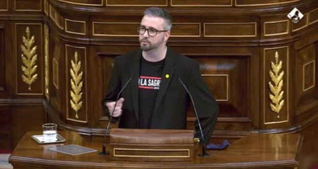 Ein Mann in einem schwarzen Blazer und Brille steht an einem Podium mit zwei Mikrofonen, einem Glas Wasser, einem Buch und einem blauen Tuch und hält eine Rede vor einer hölzernen Wand.