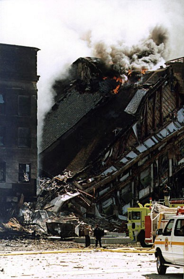 Feuerwehrfahrzeug vor einem brennenden Gebäude geparkt, mit Rauch aus Fenstern, Menschen in der Nähe und sichtbarem Himmel im Hintergrund.