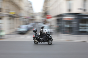 Eine Person in schwarzer Kleidung und Helm auf einem Motorroller die Straße entlangfahren, mit anderen Fahrzeugen, Gebäuden und einem klaren blauen Himmel im Hintergrund.