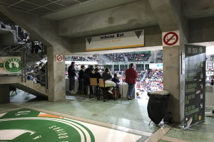 Eine Gruppe von Menschen, die vor einer sitzenden Menge in einem Stadion stehen, mit Schildern, die "Boston Celtics vs New York Jets" lesen, einem Müllcontainer, einer Tafel mit Text, einer Treppe mit Geländern und Deckenleuchten.
