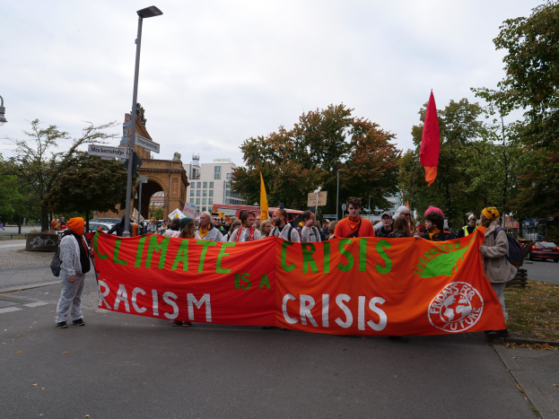 Eine Gruppe von Menschen hält ein 'Klimakrise ist eine Krise'-Schild und geht eine von Bäumen gesäumte Straße mit Fahrzeugen, Gebäuden und einem klaren blauen Himmel im Hintergrund entlang.