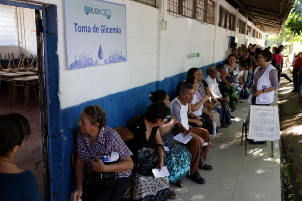 Eine Gruppe von Menschen sitzt auf Bänken vor einem Gebäude und wartet in einer Schlange, um an einer Wahllokal in Guadalajara, Mexiko, mit einer Texttafel an der Wand und einer Tür links und Bäumen im Hintergrund zu wählen.