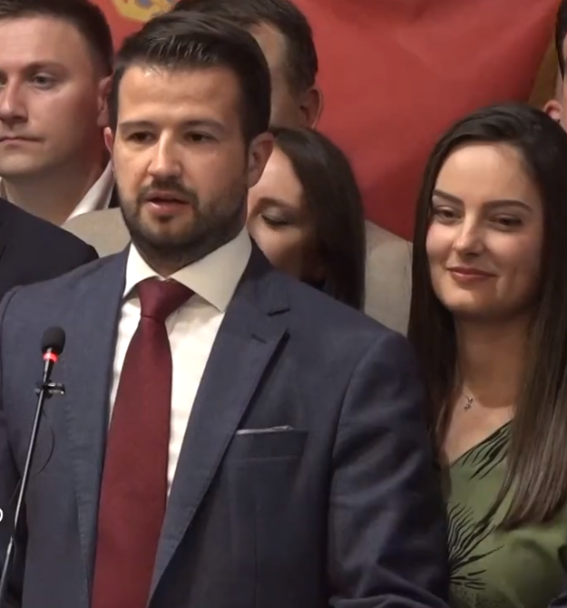 Ein Mann im Anzug und Krawatte steht an einem Rednerpult mit einem Mikrofon und einer Fahne im Hintergrund, wahrscheinlich reagiert er auf ein Regierungsverbot von sozialen Medien.