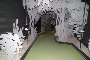 Miniatur-Golfplatz mit grüner Auslegeware, Bäumen im Hintergrund, Ständen mit Gegenständen auf beiden Seiten und Wänden mit kunstvollen Verzierungen.