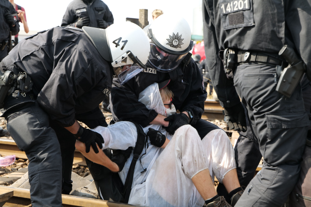 Eine Gruppe von Polizisten in Einsatzkleidung verhaftet einen Mann in einem weißen Kleid auf einem Gleis, mit anderen im Hintergrund.