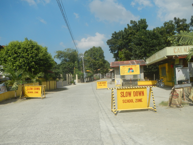 Gelbes "Slow Down School Zone"-Schild auf der linken Seite einer Straße mit geparkten Fahrzeugen auf der rechten Seite, Bäumen, Gebäuden, Strommasten, Drähten und Himmel im Hintergrund.