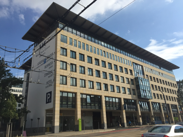 Großes modernes Gebäude mit zahlreichen Fenstern, beschriftet mit "Stadthalle Center für Unternehmen", umgeben von Bäumen und städtischer Infrastruktur in Köln, Deutschland.