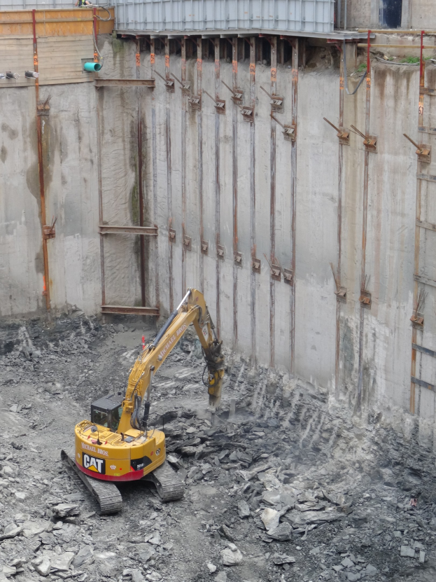 Bagger bei der Arbeit auf einer Baustelle mit Steinen und Eisenstangen, mit einem teilweise errichteten Gebäude links im Bild.