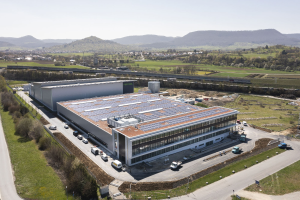 Luftbild einer großen Fabrik mit Solarmodulen auf dem Dach, umgeben von Fahrzeugen, Bäumen, Gras und Strommasten, mit Hügeln und einem klaren blauen Himmel im Hintergrund.