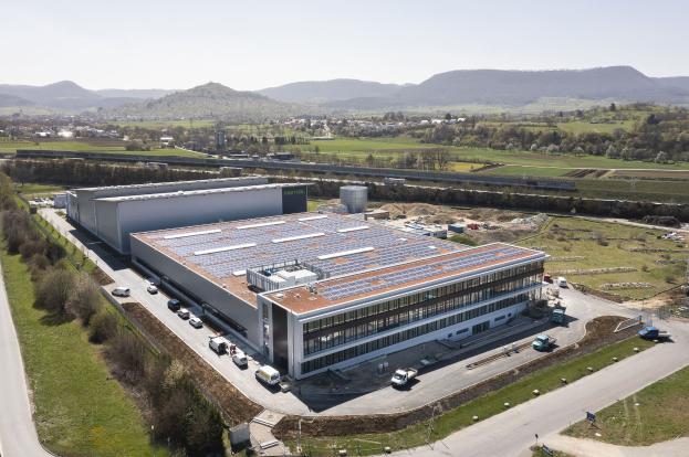 Luftbild einer großen Fabrik mit Solarmodulen auf dem Dach, umgeben von Fahrzeugen, Bäumen, Gras und Strommasten, mit Hügeln und einem klaren blauen Himmel im Hintergrund.
