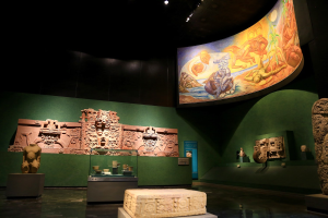 Ein Museumsausstellungsraum mit verschiedenen Artefakten, darunter Skulpturen, Tischgegenstände, eine Texttafel, eine Wandmalerei und Deckenleuchten im Mexiko-Stadt-Museum der Anthropologie.