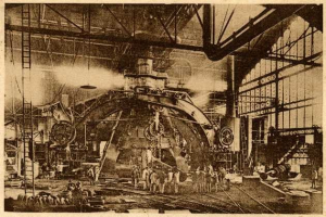 Schwarz-weiß-Foto einer Fabrik mit Arbeitern, die an einer großen Maschine arbeiten, umgeben von Leitern und Stangen, beschriftet mit "Cadillac Steel Works" unten.