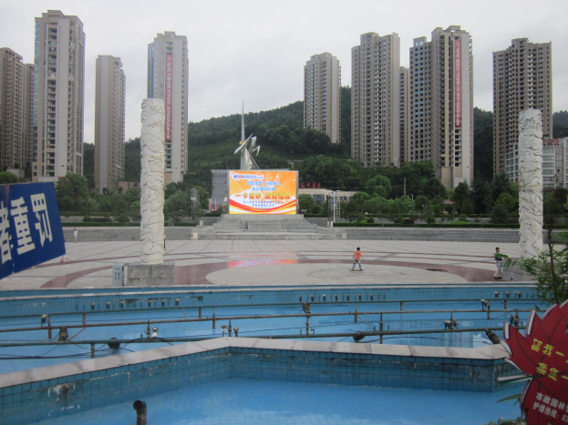 Großer Swimmingpool umgeben von hohen Gebäuden in einer städtischen Umgebung, mit wenigen Menschen in der Nähe, einem Banner auf der linken Seite, Bäumen im Hintergrund und einem klaren blauen Himmel.