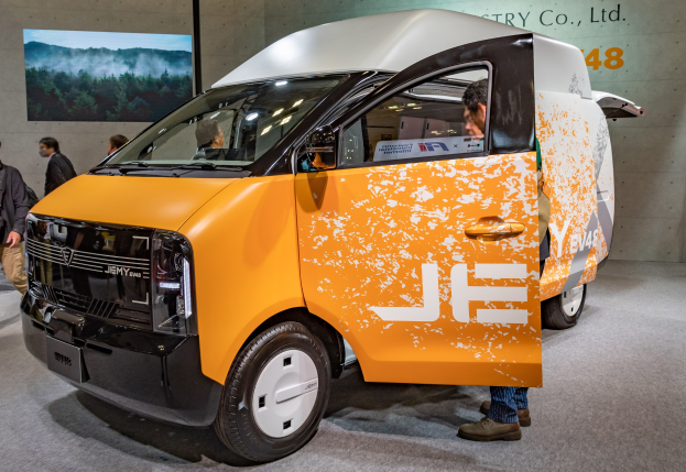 Orange und weißer Elektro-Transporter auf einer Ausstellung, umgeben von Menschen in bunten Kleidern, mit einer Wandmontage und Text im Hintergrund.