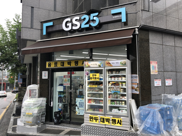 Außenansicht eines GS25-Kiosks mit Fahrzeugen auf der Straße, Bäumen und einem klaren blauen Himmel, mit Kühlgeräten, die mit Lebensmitteln gefüllt sind.