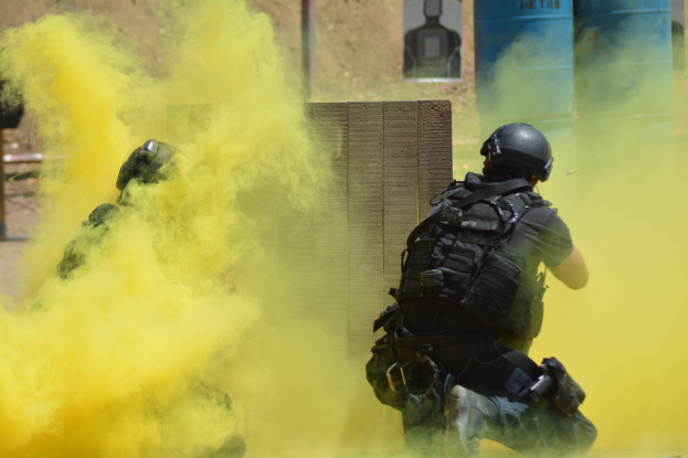 Zwei Polizisten in Einsatzkleidung mit gelbem Rauch aus dem Mund, die an einer Wand mit Text und anderen Gegenständen im Hintergrund stehen.