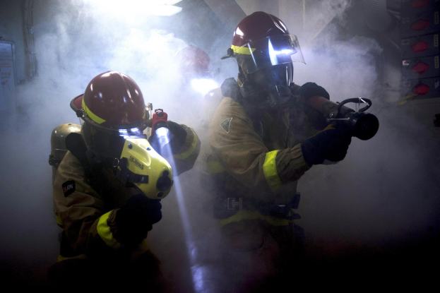 Zwei Feuerwehrleute in Schutzausrüstung sprühen Wasser auf eine rauchgefüllte Wand, mit einer Tafel und verschiedenen Gegenständen in der Nähe.