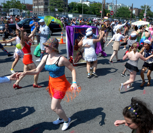 Eine Gruppe von Menschen, die eine Straße entlangmarschieren, einige mit Mötzen und Schirmen, andere tanzen, mit einer Fahne, Verkehrskegeln, Fahrzeugen, Lichtmästängen, Schildern, Bäumen, Gebäuden und einem klaren blauen Himmel im Hintergrund.