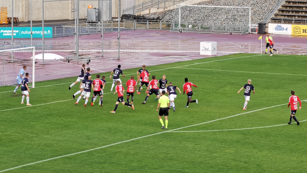 Gruppe von Menschen, die Fußball auf einem Feld mit Zuschauern in einem Stadion spielen, umgeben von Pfosten, Tafeln mit Text, Geländern, Stufen und anderen Gegenständen.