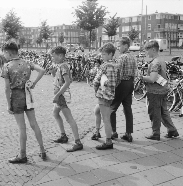 Schwarz-weiß-Foto einer Gruppe junger Jungs, die zusammen auf einem Bürgersteig stehen, umgeben von Fahrrädern, Fahrzeugen, Bäumen, Pfählen und Gebäuden im Hintergrund.