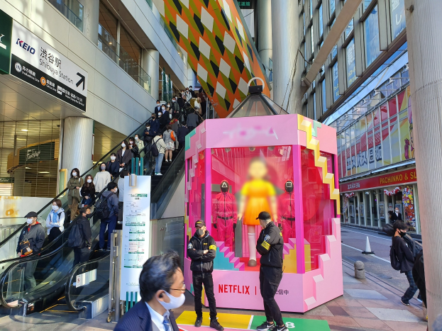 Eine Gruppe von Menschen steht vor einem Gebäude mit Glaswänden, Säulen und Texttafeln, einige tragen Masken, während eine Rolltreppe mit Menschen daneben aufsteigt; eine pinkfarbene Box in der Mitte zeigt Schaufensterpuppen und trägt die Aufschrift "Netflix's new Netflix booth in Tokyo."