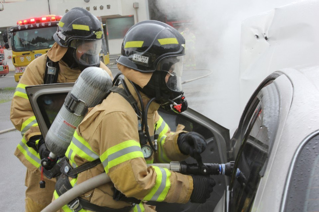 Zwei Feuerwehrleute in Schutzausrüstung verwenden einen Schlauch, um ein brennendes Auto zu löschen, während Fahrzeuge und ein Gebäude im Hintergrund zu sehen sind.