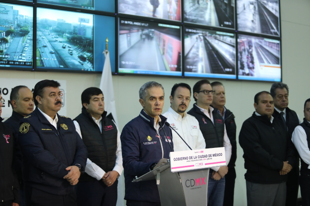 Eine Gruppe von Männern vor einem Rednerpult mit einem Mikrofon und einer Tafel, auf der "Mexikanische Zoll- und Grenzschutzbehörde" steht, mit Fernsehern und einer Flagge an der Wand dahinter.