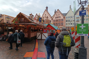 Eine Gruppe von Menschen, die auf einem Kopfsteinpflasterweg neben einem Weihnachtsmarkt in Nürnberg, Deutschland, entlanggeht, mit Laternenmasten und Texttafeln, Gebäuden mit Fenstern im Hintergrund und einem bewölkten Himmel.