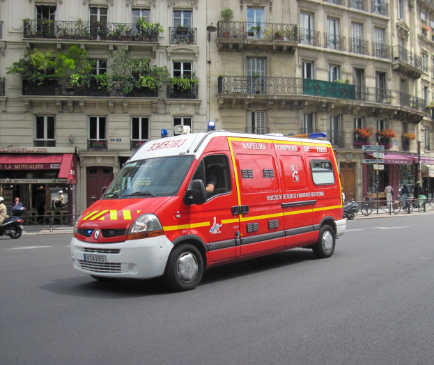 Ambulanz steht auf der Straße vor einem Gebäude mit Fenstern, Geländern, Pflanzen und anderen Gegenständen, mit einigen Menschen und Fahrrädern im Hintergrund.