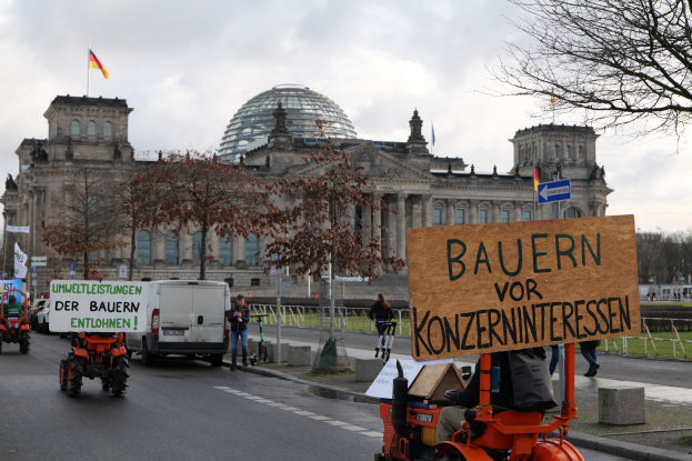 Eine Gruppe von Menschen auf Traktoren auf einer Straße vor dem Reichstaggebäude in Berlin, die Schilder halten und Bäume, Pfosten, Flaggen und Geländer passieren, unter einem bewölkten Himmel.