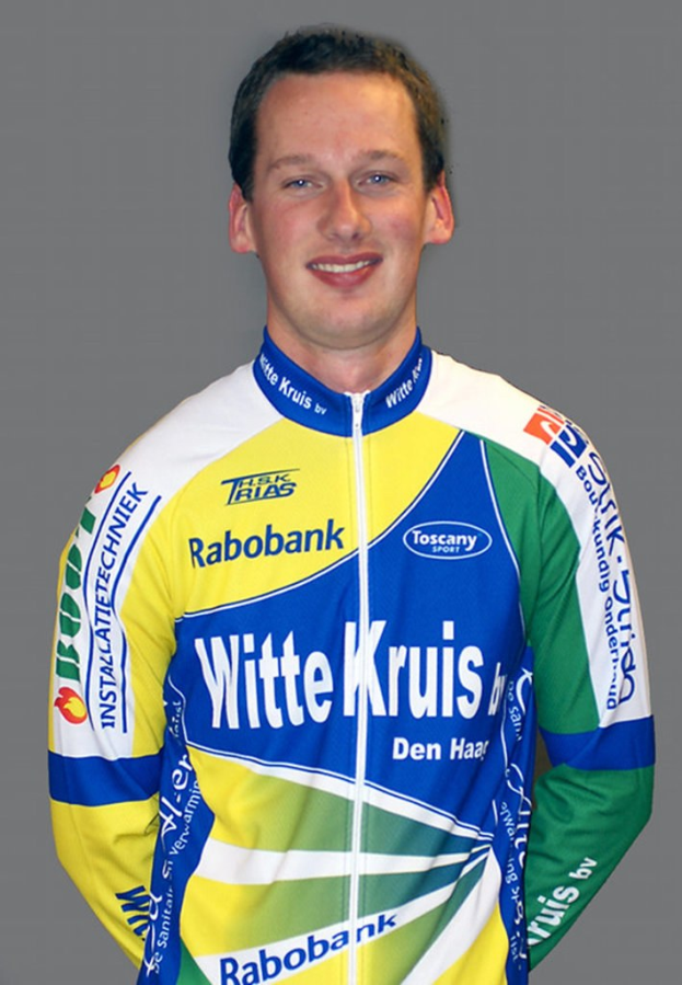 Ein männlicher Athlet posiert für ein Foto, wobei er ein Hemd trägt, auf dem mehrere Sponsorenlogos zu sehen sind.