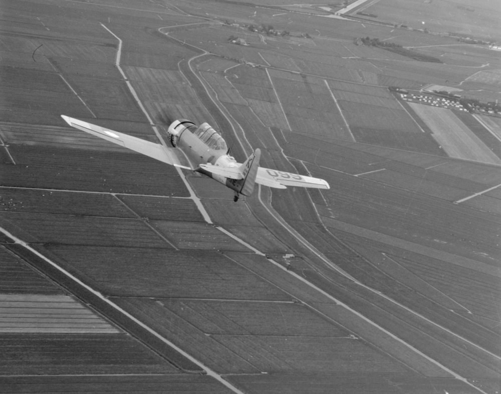 Schwarzes und weißes Bild eines kleinen Flugzeugs, das über ein Feld mit Kulturpflanzen fliegt und mittig im Bild platziert ist.