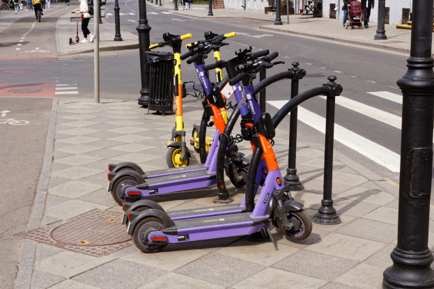 Eine Reihe von Elektro-Scootern, die entlang einer Straße geparkt sind, mit Fußgängern und Fahrradfahrern auf dem Gehweg, einem Müllcontainer im Vordergrund und Gebäuden, Bäumen und Pflanzen im Hintergrund.