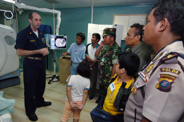 Eine Gruppe von Menschen in einem Krankenhauszimmer mit einem Mann, der ein Mikrofon hält, ein drapiertes Bett, medizinische Geräte, ein Röntgengerät, eine Texttafel, eine Tür und Deckenleuchten.