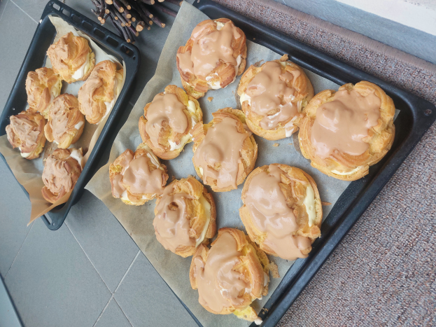 Ein Tablett mit verschiedenen Danish-Pasteten in hellgoldbrauner Farbe und flaky Textur, einige mit Puderzucker bestäubt, liegt auf einer Theke neben einem offenen Fenster und wird von warmem Licht durchflutet.