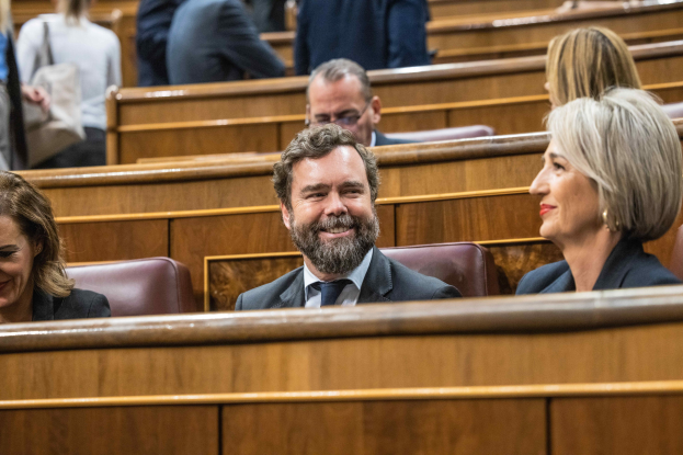 Eine Gruppe von Menschen, die in einem Gerichtssaal sitzen, mit einem Mann in Anzug und Krawatte und zwei leger gekleideten Frauen, die nebeneinander sitzen, sowie Bänke im Hintergrund.