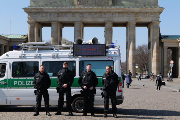 Polizisten in schwarzen Uniformen stehen vor dem Brandenburger Tor in Berlin, mit einem weißen und grünen Fahrzeug im Vordergrund und Fußgängern und Radfahrern im Hintergrund.