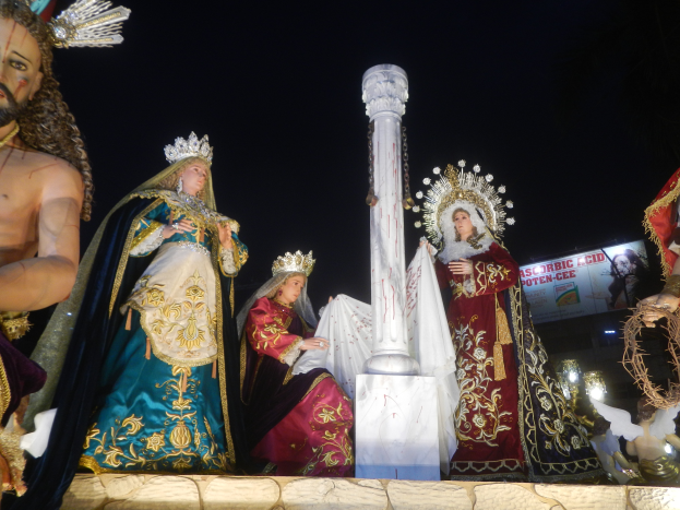Eine Gruppe von beleuchteten Jesus- und Marienstatuen auf einem Paradewagen, umgeben von einer Säule und einem Banner mit Text, vor einem dunklen Hintergrund.